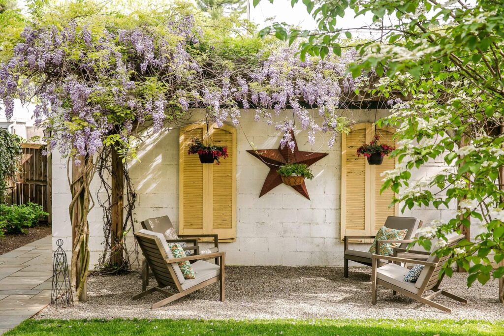 A peaceful outdoor setting with two cushioned wooden chairs on a gravel patio, framed by a white brick wall with yellow shutters, a central star-shaped decor, and hanging baskets of purple wisteria under a pergola.