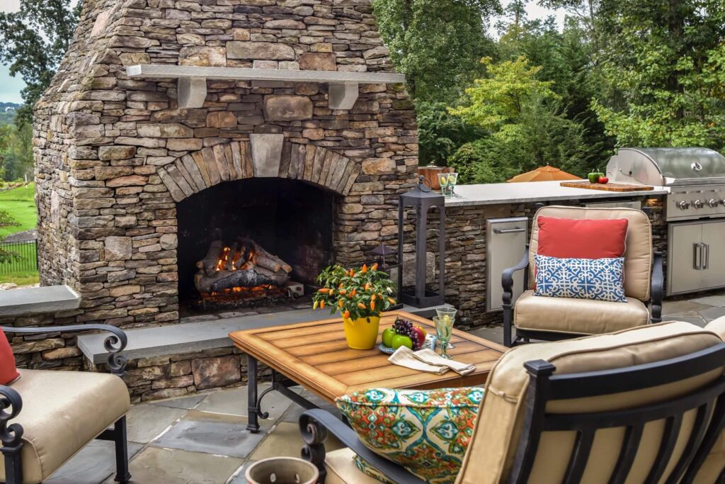 A stone patio featuring a lit outdoor fireplace, a built-in grill, and a table with food and drinks, surrounded by cushioned chairs and dense greenery.