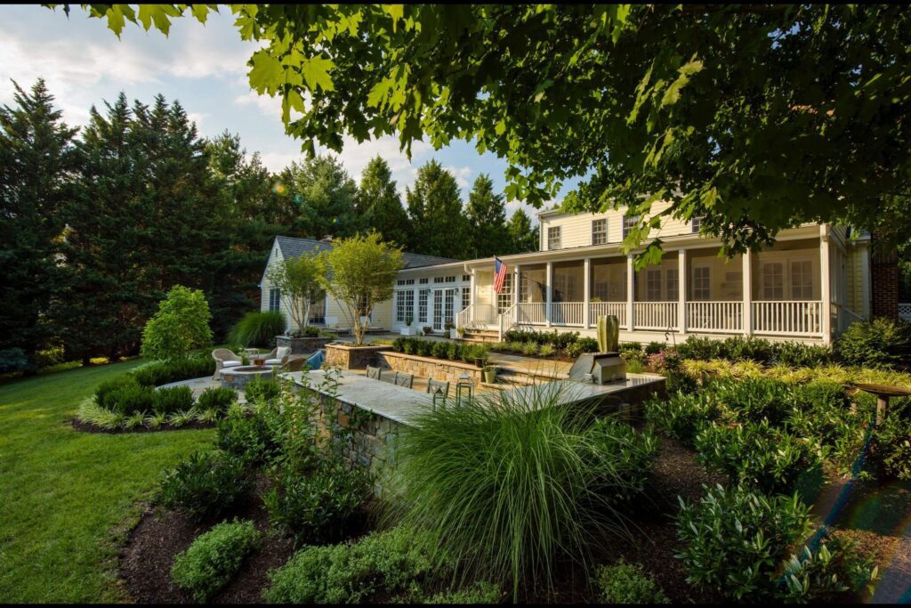 A lush backyard with stone patio seating and a built-in bar, set against a white house with an American flag, surrounded by trees and manicured gardens.